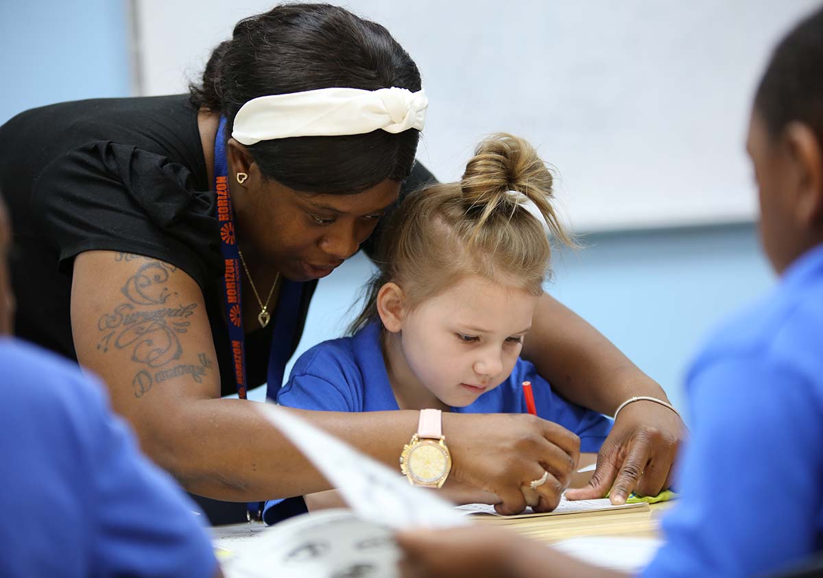 HSA Teacher smiles while kneeling beside a young student in a classroom setting.